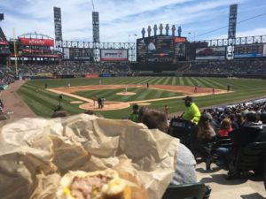 Photo of Bratwurst in foreground, White Sox Park, from behind home plate on the first base side, in the background. 