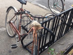 Photo of barbie dolls attached to handlebar bike basket.